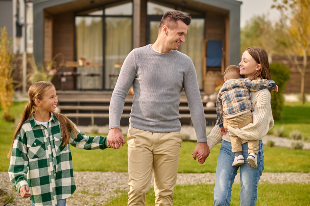 Family, togetherness. Woman with child man and girl of school age holding hands together happy looking at each other walking in courtyard of country house on fine day